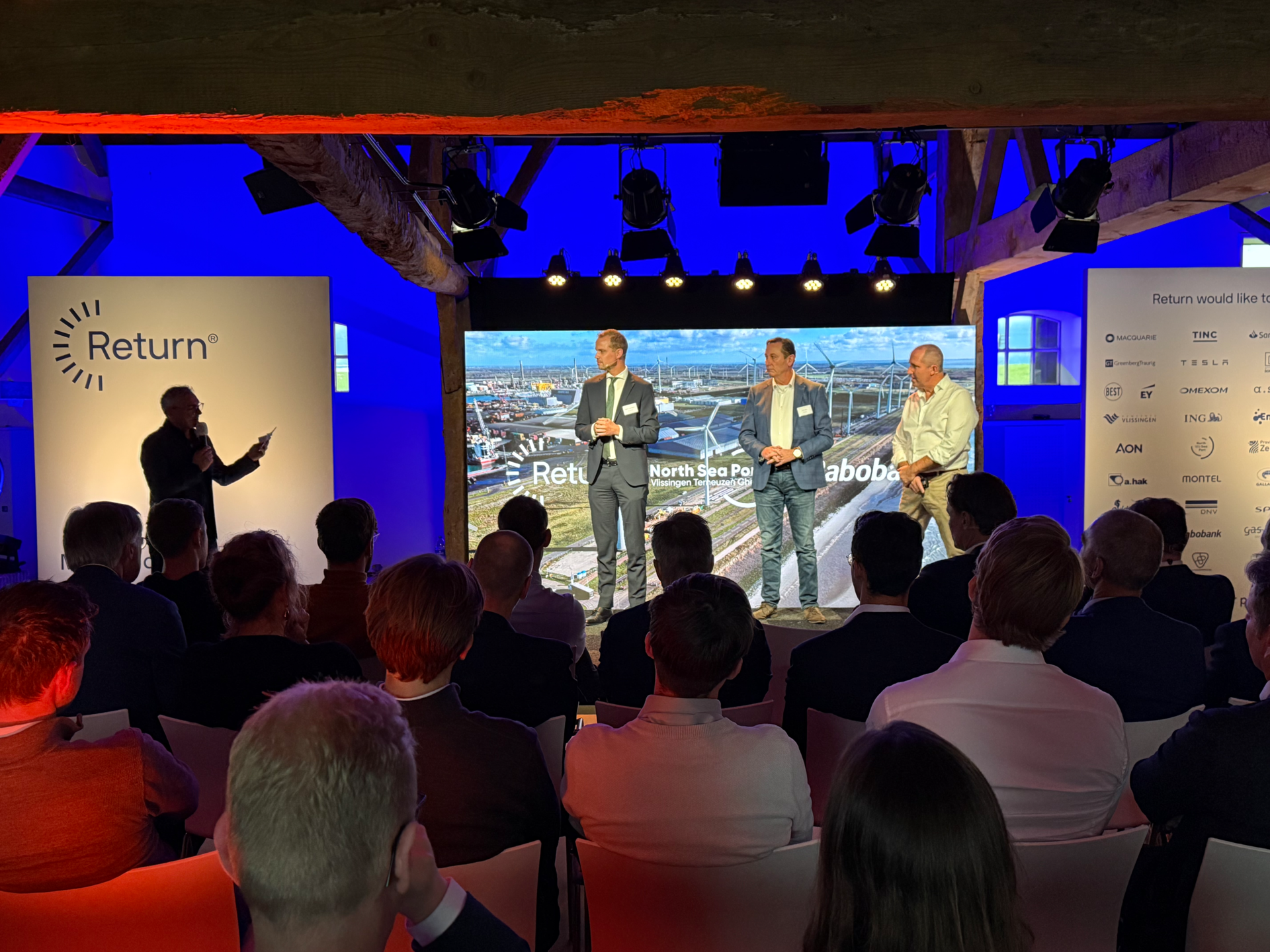 Photo of speakers on stage at Mufasa event with Return and North Sea Port, audience and harbour screen image.